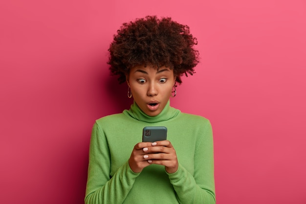 Бесплатное фото Концепция технологий и эмоций. ошеломленная темнокожая женщина пользуется современным мобильным телефоном, смотрит на дисплей, удивлена хорошим гаджетом, болтает в сети.