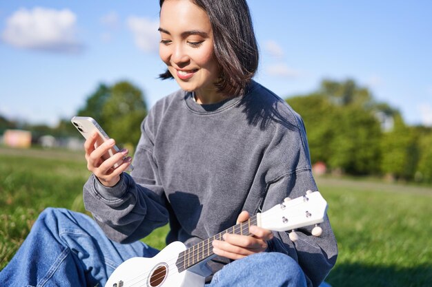 無料写真 技術と音楽スマート フォンで公園に座って、ウクレレを保持している笑顔のアジアの流行に敏感な女の子