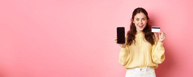 Бесплатное фото Технологии и онлайн-покупки молодая привлекательная модель, показывающая пустой экран смартфона с пластиковой кредитной картой, показывает банковский счет или приложение, стоящее на розовом фоне