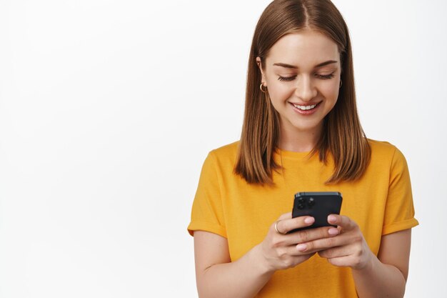 Technology and cellular. Smiling young woman looking at phone screen, using cellphone, smartphone application, standing happy against white background