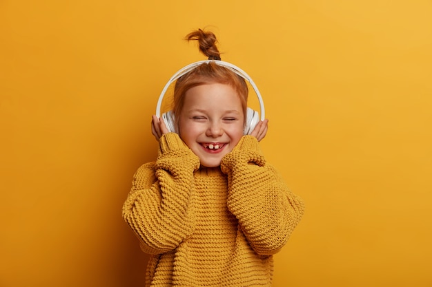 テクノロジー、子供、音楽のコンセプト。生姜髪のかわいい笑顔の小さな子供はステレオヘッドホンを着用し、純粋な音を楽しみ、好きな歌を聴き、楽しく笑い、ニットのセーターを着ています