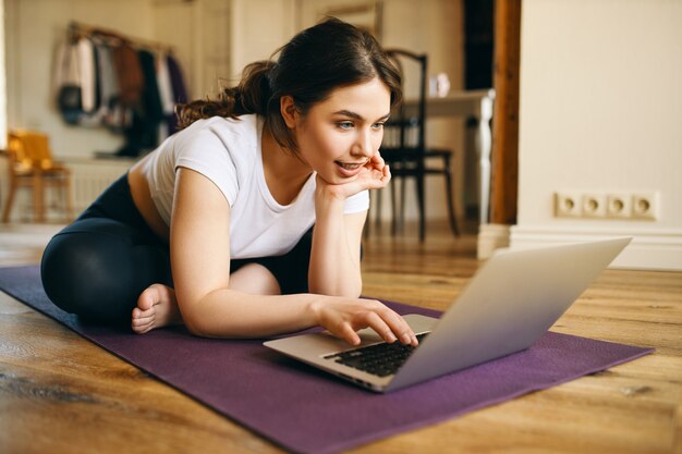 テクノロジー、コミュニケーション、遠隔教育、社会的距離。ノートパソコンでワイヤレス高速インターネット接続を使用して、オンラインでヨガインストラクターコースを見て、マットの上に座っているかわいいプラスサイズの女の子