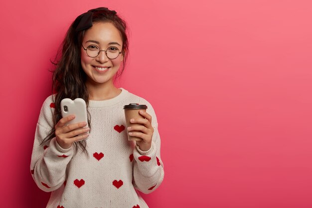 Technology, communication and lifestyle concept. Pretty young girl in round glasses uses smartphone for reading cool blog and messaging, drinks takeaway coffee, downloads app, stands indoor.