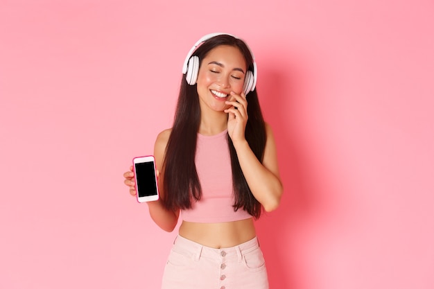 技術、コミュニケーション、オンラインライフスタイルのコンセプト。ヘッドフォンで素敵な夢のようなアジアの女の子、目を閉じて、愚かな頬に触れる、音楽を聴いている間に空想、モバイル画面を表示