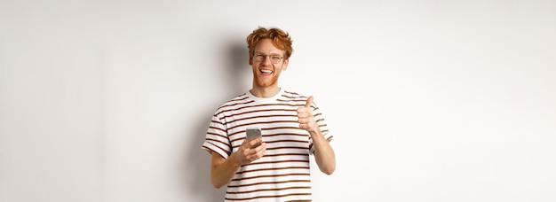 Free photo technology and ecommerce concept satisfied male model with red hair showing thumbsup and holding sma