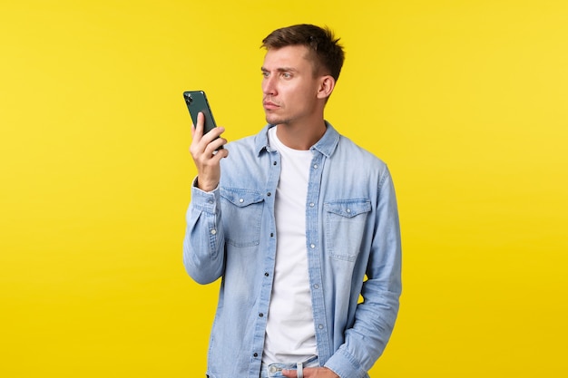 Концепция технологии, образа жизни и рекламы. Раздраженный и разочарованный молодой человек недоумевает по поводу внезапно закончившейся беседы, раздраженно смотрит в мобильный телефон, стоя на желтом фоне.