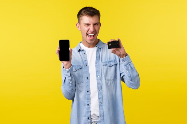 Technology, lifestyle and advertisement concept. Enthusiatic happy blond guy showing credit card and mobile phone, smiling and saying yes pleased, bought cool thing on sale online, yellow background.