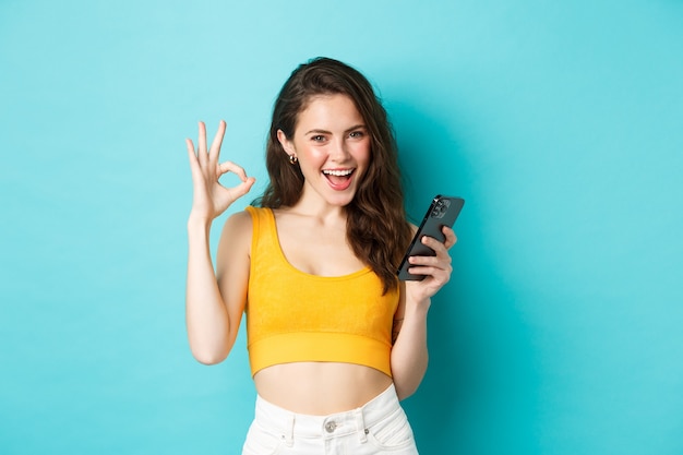Концепция технологии и образа жизни. Уверенно привлекательная женщина говорит «да», показывая знак «хорошо» к чему-то в Интернете, держа смартфон в руках, стоя на синем фоне.