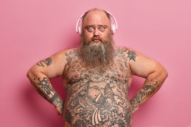 Free photo technology and lifestyle concept. serious confident plump man wears headphones, listens music, has tattooed body, fat stomach, thick beard, poses against pink wall, found great playlist