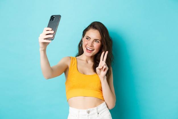 Technology and lifestyle concept. Stylish young woman taking selfie in her summer clothes, showing v-sign at smartphone camera, standing over blue background