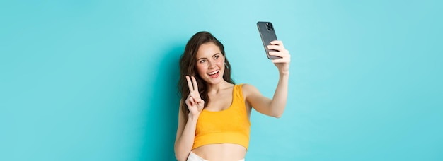 Technology and lifestyle concept stylish young woman taking selfie in her summer clothes showing vsi