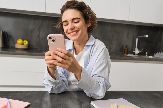 Technology and lifestyle young woman sits at home uses smartphone in her kitchen and smiles