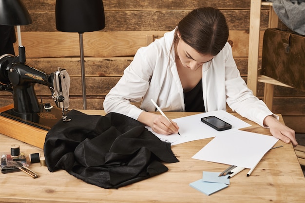 Free photo technology makes work easier. portrait of focused determined designer drawing new garment concept, measure everything and calculating with smartphone, sitting near sewing machine and fabric