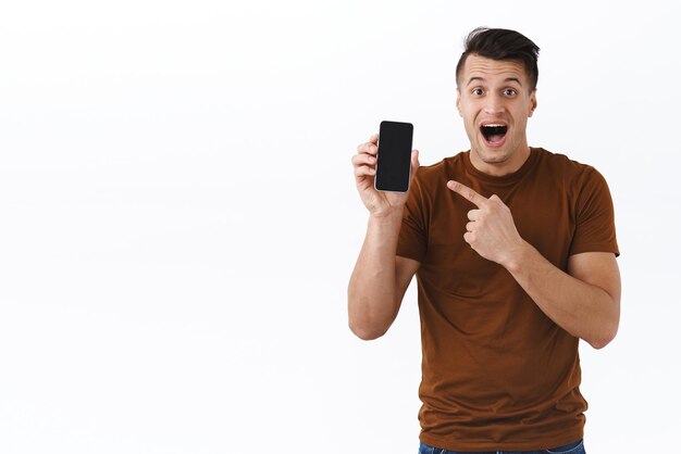 Технологии онлайн-образа жизни и концепции общения Взволнованный и пораженный кавказский парень, указывающий пальцем на мобильный телефон, впечатлен открытым ртом, представляя новое приложение для интернет-магазина