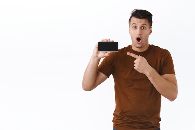 Технологии онлайн образа жизни и концепция коммуникации Шокированный и взволнованный красивый кавказский мужчина с открытым ртом, указывающий пальцем на экран мобильного телефона, представляя сотовый продукт приложения