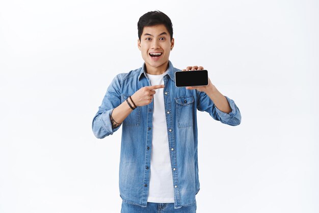 Technology online lifestyle concept Must have for your entertainment Portrait of cheerful young asian man showing application or video on smartphone display pointing mobile screen smiling