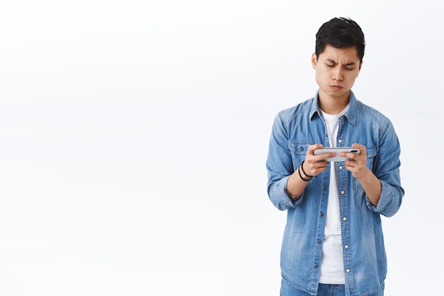 Technology online lifestyle concept Portrait of bothered and annoyed young gloomy asian guy cant win in game look upset losing level holding mobile phone both hands frowning at screen