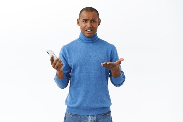 Technology online lifestyle concept Portrait of displeased young africanamerican man shrugging in dismay being disappointed holding mobile phone read strange frustrating news internet