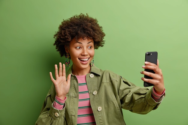 technology and online lifestyle concept. smiling woman says hi enjoys video calling uses high speed internet connection dressed in fashionable jacket isolated on green wall