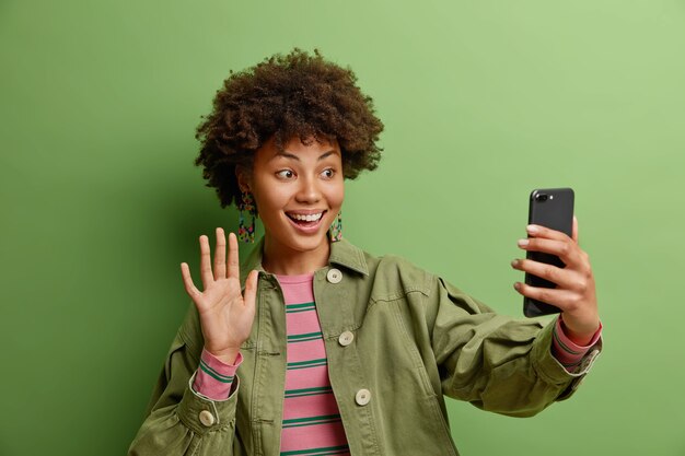 Technology and online lifestyle concept. Smiling woman says hi enjoys video calling uses high speed internet connection dressed in fashionable jacket isolated on green wall