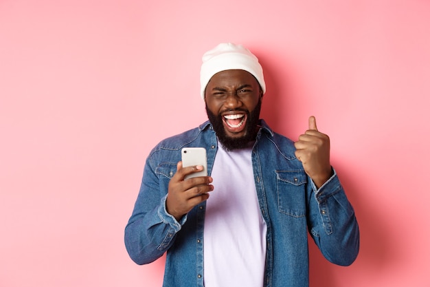 Technology and online shopping concept. Happy Black man rejoicing, winning in app, holding smartphone and scream yes, standing over pink background