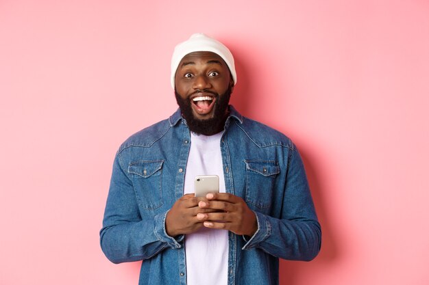 Технологии и концепция покупок в Интернете. Удивленный молодой темнокожий мужчина с помощью мобильного телефона, глядя в камеру, изумлен и счастлив после прочтения сообщения, розовый фон