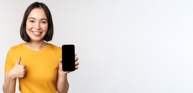 기술 및 사람 개념 흰색 배경에 서 있는 스마트폰 화면 휴대 전화 앱 인터페이스와 엄지손가락을 보여주는 웃는 젊은 여성