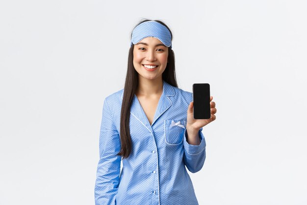기술, 사람 및 가정 레저 개념입니다. 잠옷을 입고 행복한 미소를 짓고 있는 아시아 소녀와 수면 마스크는 모바일 앱으로 그녀의 수면을 추적하고 스마트폰 화면을 보여주고 만족스러운 흰색 배경을 보여줍니다.