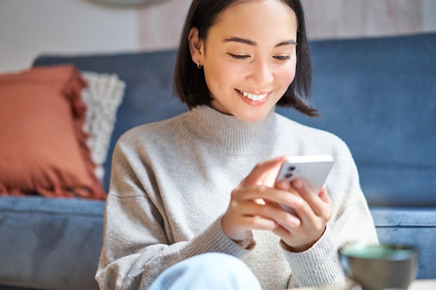 テクノロジーと人々 スタイリッシュなアジアの若い女性が家に座って、スマートフォンのテキスト メッセージを使用して