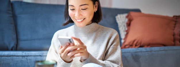 Технологии и люди Молодая стильная азиатка сидит дома с текстовым сообщением на смартфоне