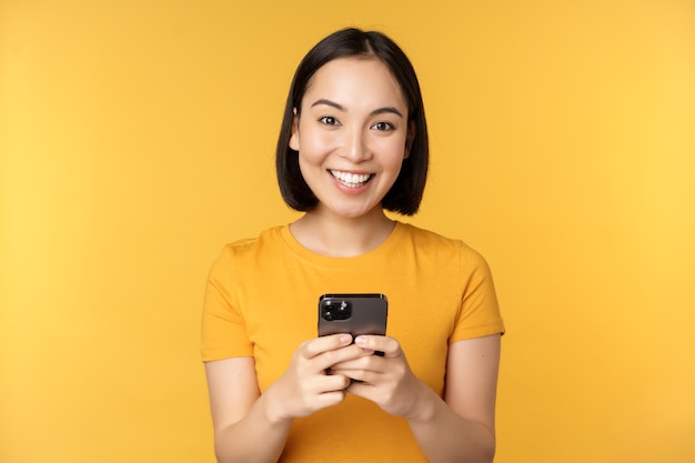 Technology Smiling asian woman using mobile phone holding smartphone in hands standing in tshirt against yellow background