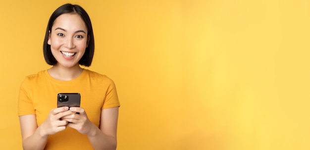 黄色の背景に対してTシャツに立っている手でスマートフォンを保持している携帯電話を使用してアジアの女性を笑顔