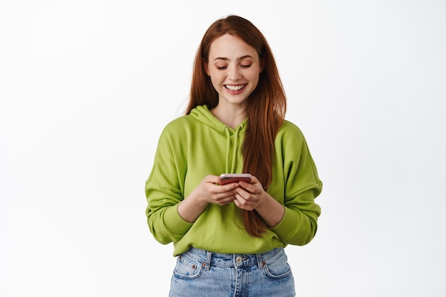 Technology. Smiling redhead girl looking at smartphone, chatting on phone, using application on cellphone, standing in hoodie against white background.