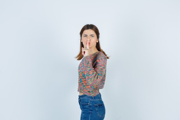 Teen beautiful girl showing silence gesture in sweater, jeans and looking annoyed , front view.