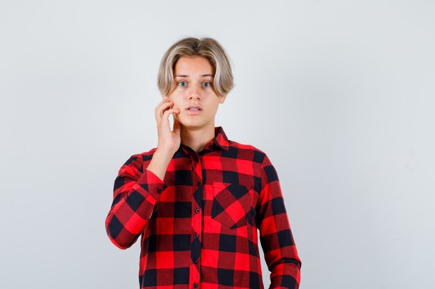 Teen blonde male keeping hand on face in casual shirt and looking surprised. front view.