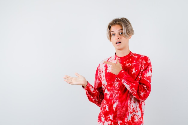 Teen blonde male in oversized shirt pointing left, showing welcome gesture and looking bewildered , front view.
