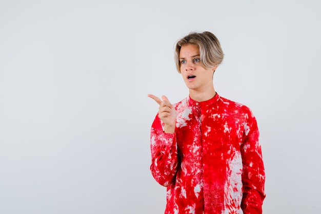 Teen blonde male pointing away in oversize shirt and looking shocked , front view.