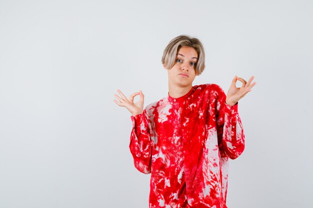 Teen blonde male showing ok gesture in oversized shirt and looking pensive. front view.