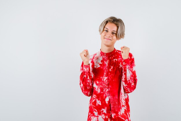 Teen blonde male showing winner gesture in oversized shirt and looking proud , front view.