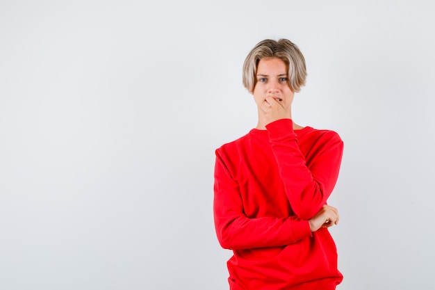 Teen boy biting his nail in red sweater and looking uneasy , front view.