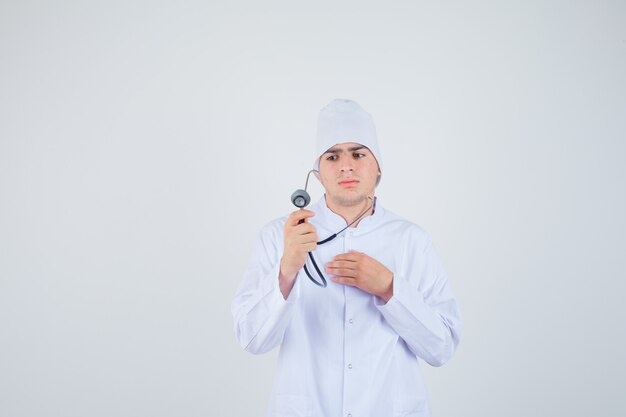 teen boy in doctor uniform holding stethoscope while pretending to check heart beat and looking serious