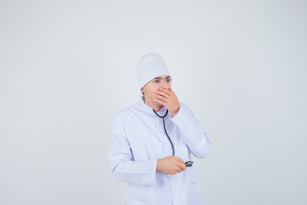 teen boy holding hand on mouth in doctor uniform and looking puzzled