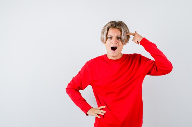 Teen boy keeping finger on temple, opening mouth in red sweater and looking furious . front view.