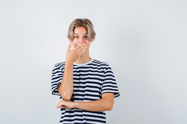 Teen boy keeping hand on mouth in t-shirt and looking amazed. front view.