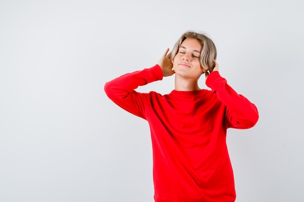 Teen boy keeping hands on head, closing eyes in red sweater and looking charming. front view.