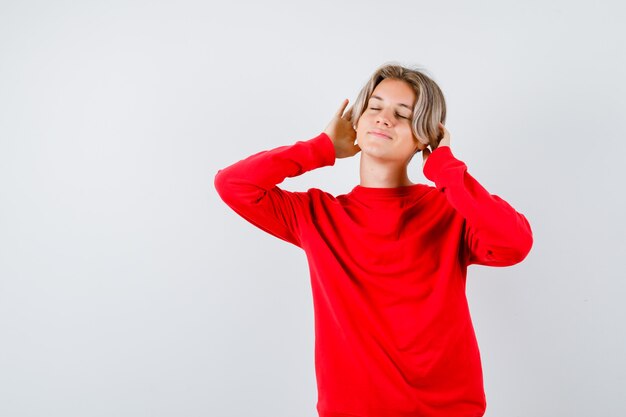 Мальчик-подросток держит руки на голове, закрывает глаза в красном свитере и выглядит очаровательно. передний план.