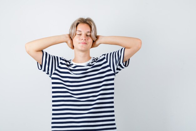 Teen boy keeping hands behind head, closing eyes in t-shirt and looking relaxed. front view.