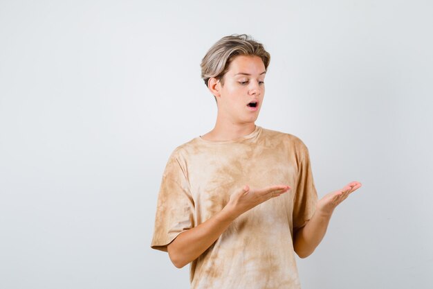 Teen boy looking at his palms, opening mouth in t-shirt and looking astonished , front view.