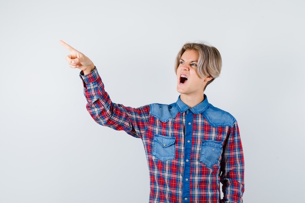 Teen boy pointing away in checkered shirt and looking annoyed , front view.