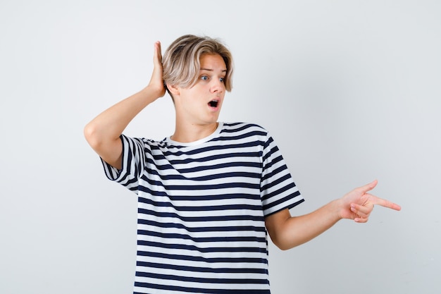 Teen boy pointing to the right side, keeping hand behind head in t-shirt and looking horrified . front view.